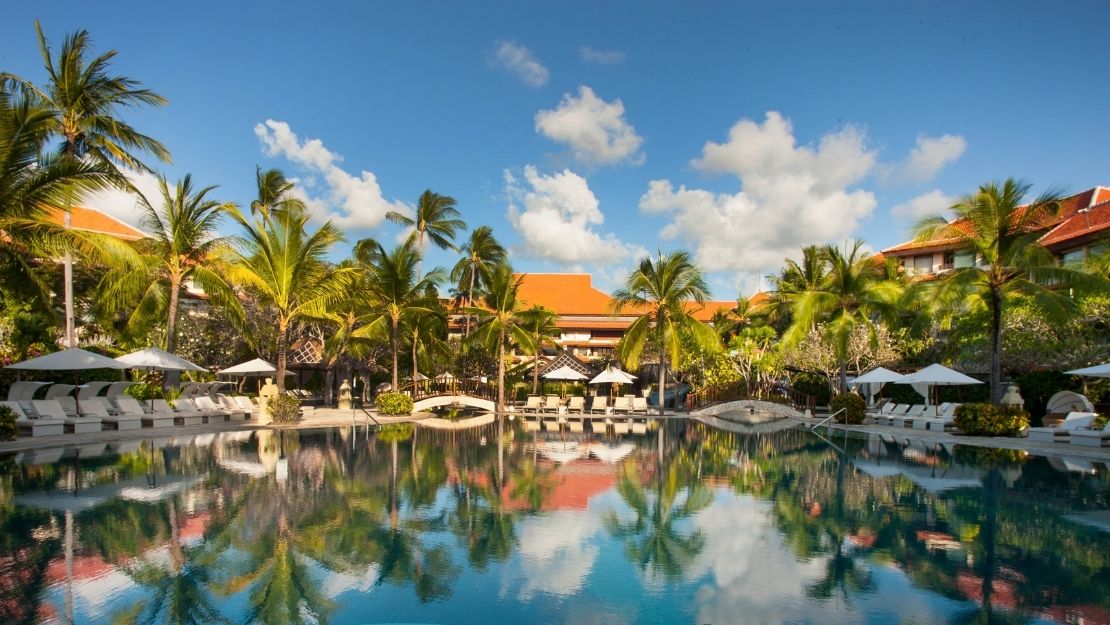Resort swimming pool surrounded by palm trees, lounge chairs, umbrellas, and red-roofed buildings under a blue sky with scattered clouds.