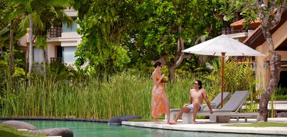 A man sits on a sun lounger by a pool under a white umbrella, wearing swim trunks. A woman in an orange sarong stands nearby. Lush greenery surrounds them.