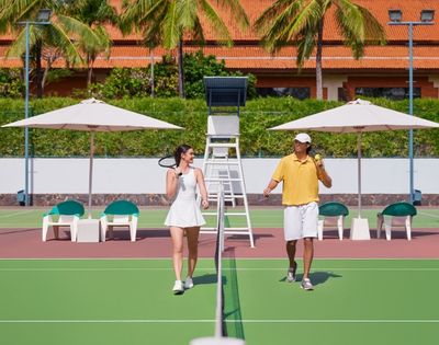 A man and woman walk on a sunny outdoor tennis court, each holding rackets, smiling. Palm trees and umbrellas create a tropical backdrop.