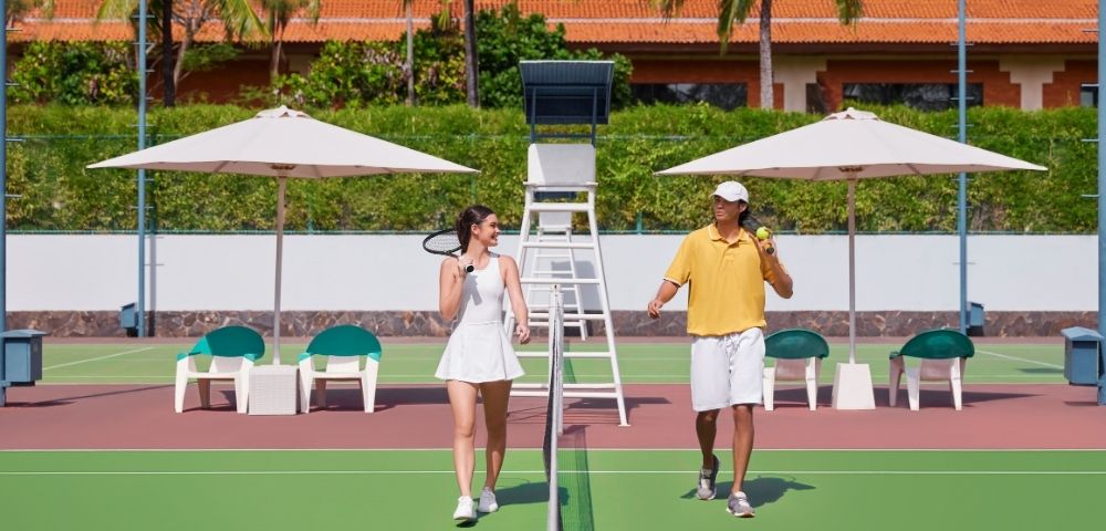 A man and woman walk on a sunny outdoor tennis court, each holding rackets, smiling. Palm trees and umbrellas create a tropical backdrop.