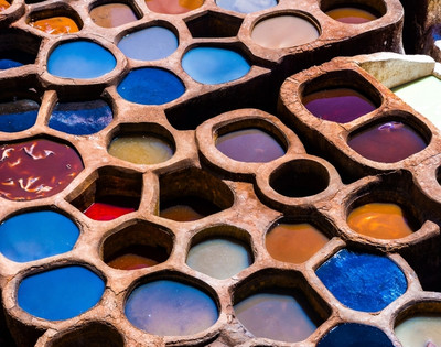 Colorful dye pits in an open-air tannery form a honeycomb pattern. Vivid blues, oranges, and reds convey a vibrant, artisanal atmosphere.