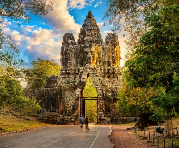Ancient stone gate with carved faces, surrounded by lush greenery, under a partly cloudy sky during golden hour, with two cyclists passing through.