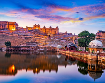 A historic fort with illuminated walls sits atop a hill, reflected in a tranquil lake during sunset. The sky is vibrant with hues of pink and orange.