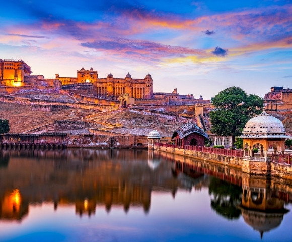 A historic fort with illuminated walls sits atop a hill, reflected in a tranquil lake during sunset. The sky is vibrant with hues of pink and orange.