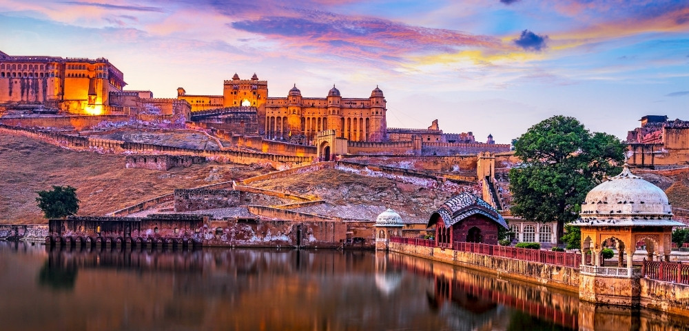 A historic fort with illuminated walls sits atop a hill, reflected in a tranquil lake during sunset. The sky is vibrant with hues of pink and orange.