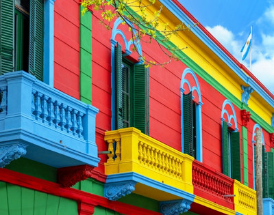 Vibrant building with red, blue, and yellow walls. Green shutters, decorative balconies. Argentine flag waves atop. Bright, cheerful atmosphere.