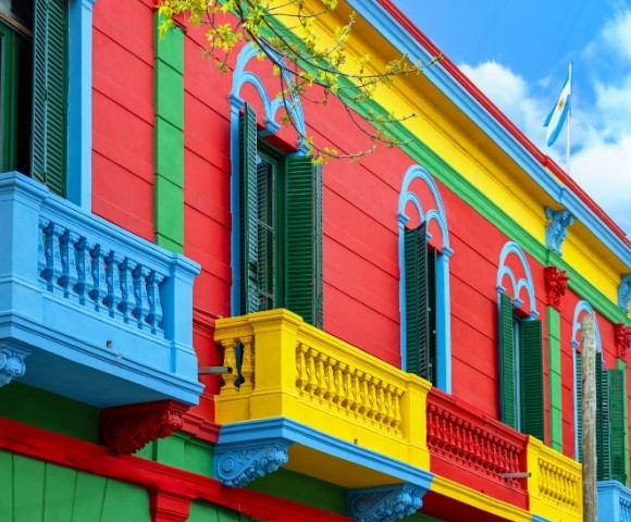 Vibrant building with red, blue, and yellow walls. Green shutters, decorative balconies. Argentine flag waves atop. Bright, cheerful atmosphere.