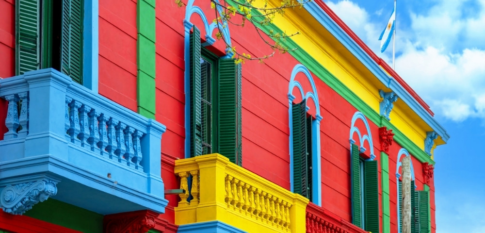 Vibrant building with red, blue, and yellow walls. Green shutters, decorative balconies. Argentine flag waves atop. Bright, cheerful atmosphere.
