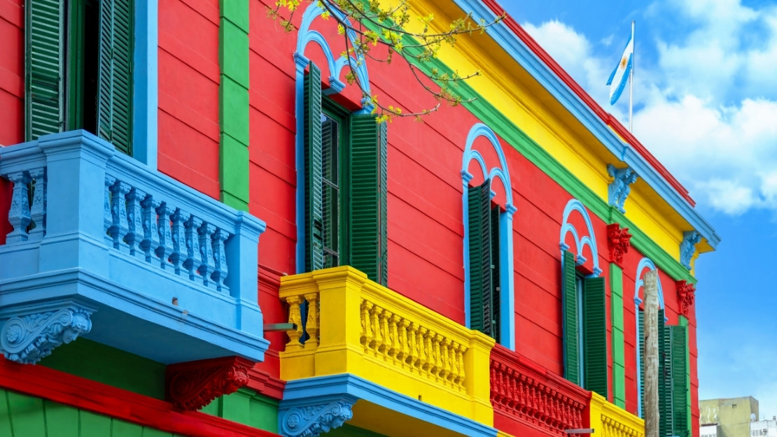 Vibrant building with red, blue, and yellow walls. Green shutters, decorative balconies. Argentine flag waves atop. Bright, cheerful atmosphere.