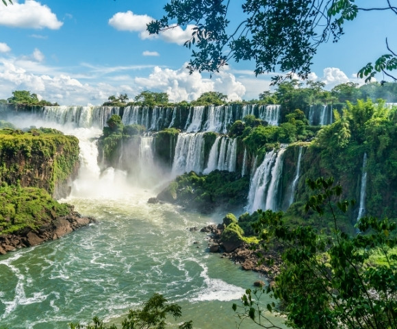 Expansive view of waterfalls cascading over lush, green cliffs into a wide, misty river under a bright blue sky, evoking awe and serenity.