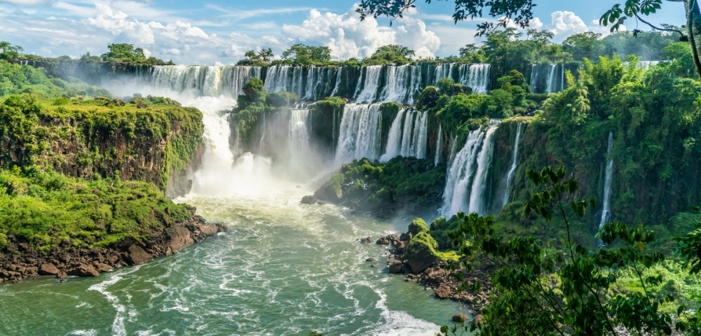 Expansive view of waterfalls cascading over lush, green cliffs into a wide, misty river under a bright blue sky, evoking awe and serenity.