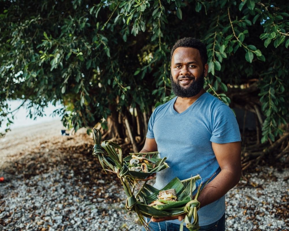 A man with a beard smiles and holds two woven leaf containers filled with food in an outdoor setting with trees and a beach in the background.