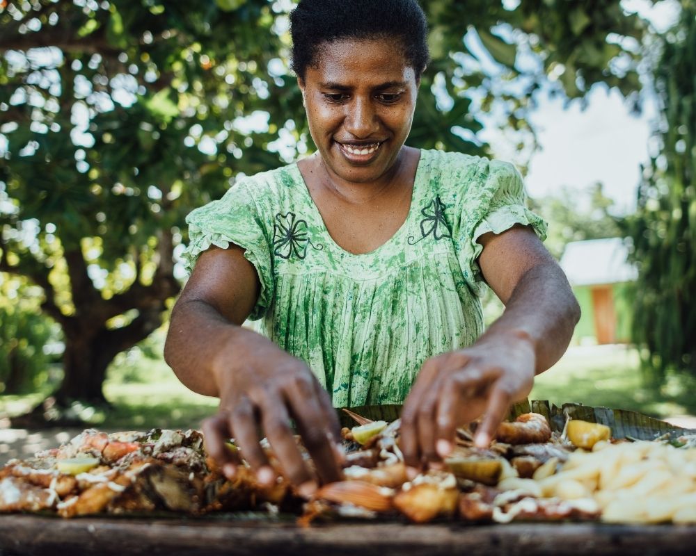 A woman in a green dress smiles as she arranges a large platter of seafood and other food items on banana leaves outdoors.
