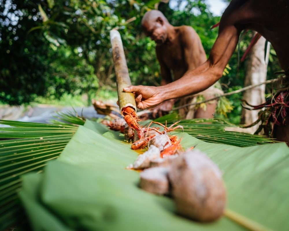 A close-up of cooked lobsters being arranged on large, green banana leaves, as a person holds a bamboo tube from which more lobsters are being placed. Another person stands behind, in a tropical, forested setting.