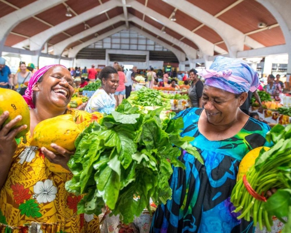 Two women laugh while holding fresh produce at an outdoor market.
