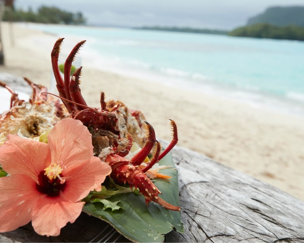 A cooked lobster garnished with a peach hibiscus flower and lettuce sits on a wooden surface with a tropical beach and turquoise ocean in the blurred background.
