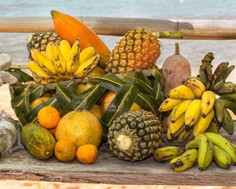 A colorful assortment of tropical fruits, including pineapples, bananas, oranges, and a papaya, is displayed in a woven basket made of palm leaves on a wooden surface with a beach and ocean in the background.