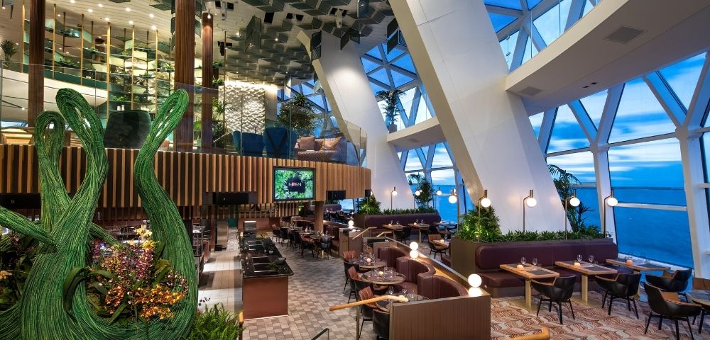 Modern restaurant interior with large geometric windows overlooking the ocean, featuring plush seating, decorative greenery sculptures, and contemporary ceiling design.