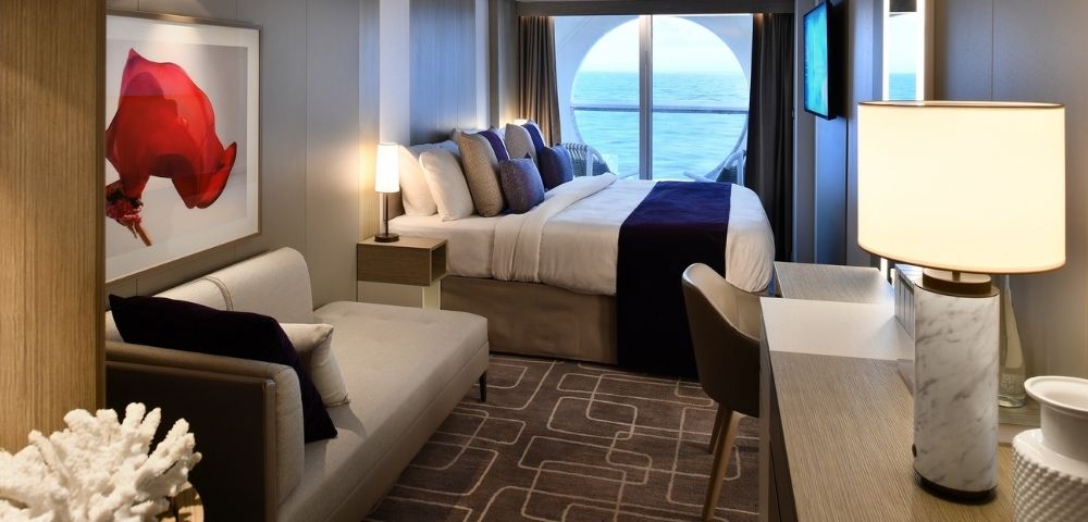 Modern cruise ship stateroom with a large bed, chaise lounge, desk, and a circular window overlooking the ocean.
