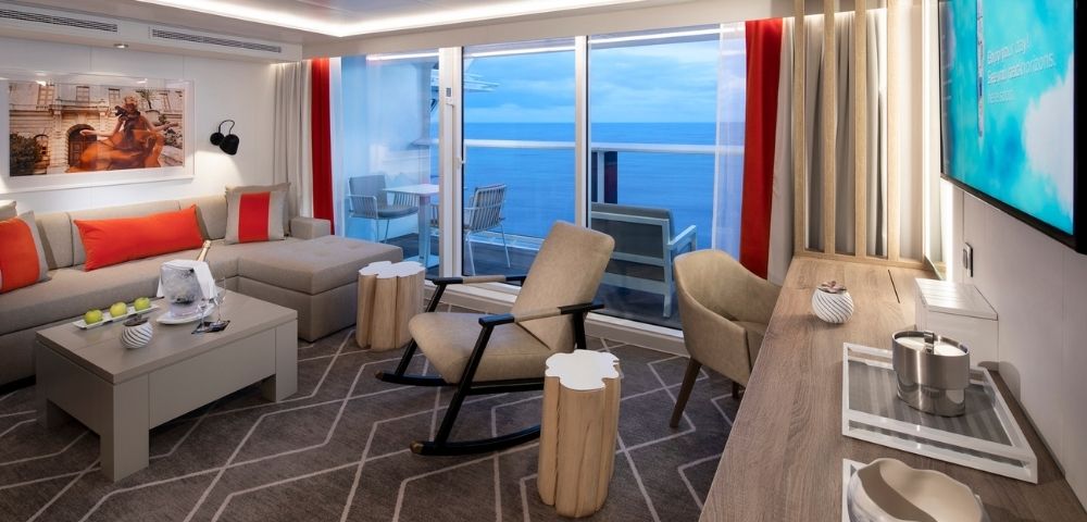 A luxurious living room with a sectional sofa, rocking chair, and a balcony overlooking the ocean on a cruise ship.