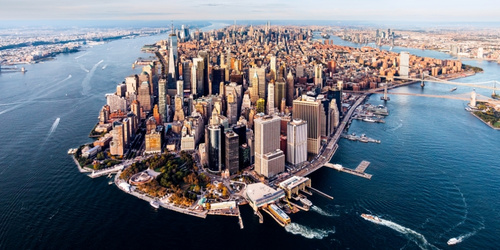 Aerial view of Manhattan, featuring dense skyscrapers surrounded by the Hudson and East Rivers. The skyline is vibrant under a blue sky.
