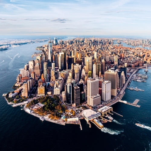 Aerial view of Manhattan, featuring dense skyscrapers surrounded by the Hudson and East Rivers. The skyline is vibrant under a blue sky.