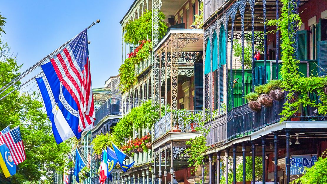 Colorful street view of New Orleans' French Quarter, featuring ornate iron balconies with lush plants. U.S. and other flags wave vibrantly.
