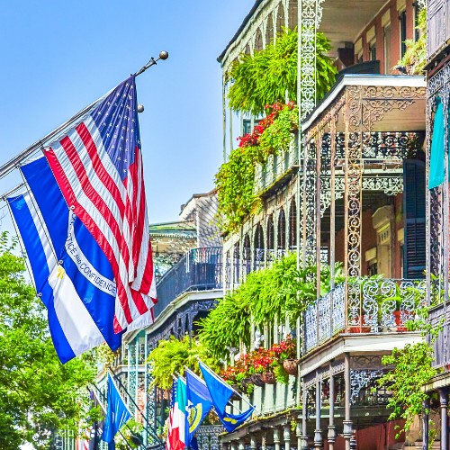 Colorful street view of New Orleans' French Quarter, featuring ornate iron balconies with lush plants. U.S. and other flags wave vibrantly.