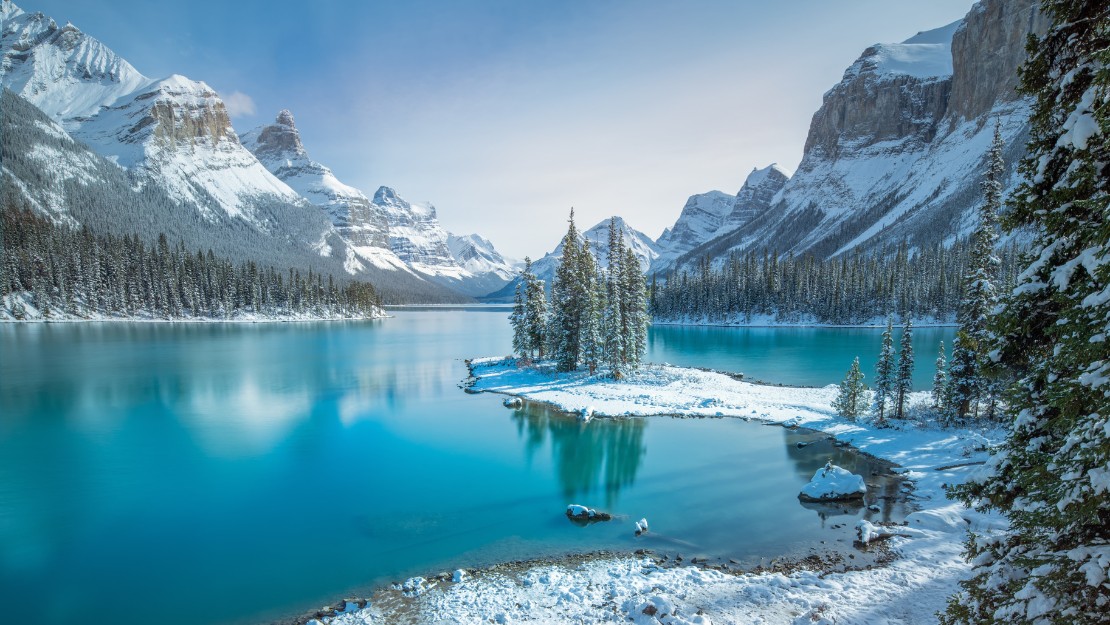 Snow-covered trees on a small island in a turquoise lake, surrounded by snow-capped mountains under a clear sky, creating a serene winter scene.