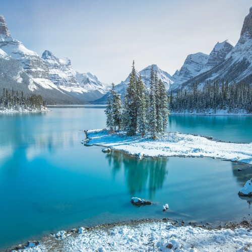 Snow-covered trees on a small island in a turquoise lake, surrounded by snow-capped mountains under a clear sky, creating a serene winter scene.