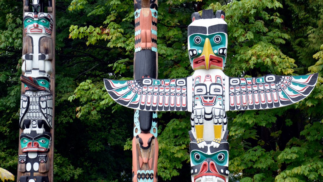 Brightly painted totem poles stand before lush green trees. Central pole features a prominent bird with outstretched wings, evoking cultural richness.