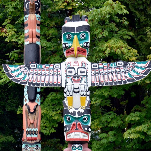 Brightly painted totem poles stand before lush green trees. Central pole features a prominent bird with outstretched wings, evoking cultural richness.