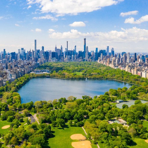 Aerial view of Central Park in New York City, featuring a large, lush, green area with a central water body. Skyscrapers line the horizon under a blue sky.