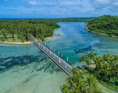 Aerial view of a modern bridge crossing a clear turquoise lagoon, surrounded by lush green palm trees and tropical foliage under a blue sky.