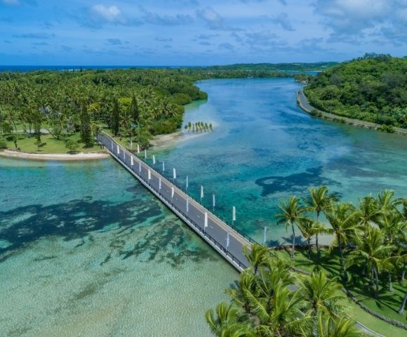 Aerial view of a modern bridge crossing a clear turquoise lagoon, surrounded by lush green palm trees and tropical foliage under a blue sky.