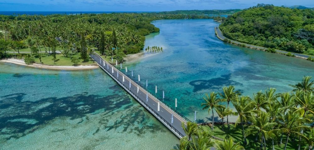 Aerial view of a modern bridge crossing a clear turquoise lagoon, surrounded by lush green palm trees and tropical foliage under a blue sky.