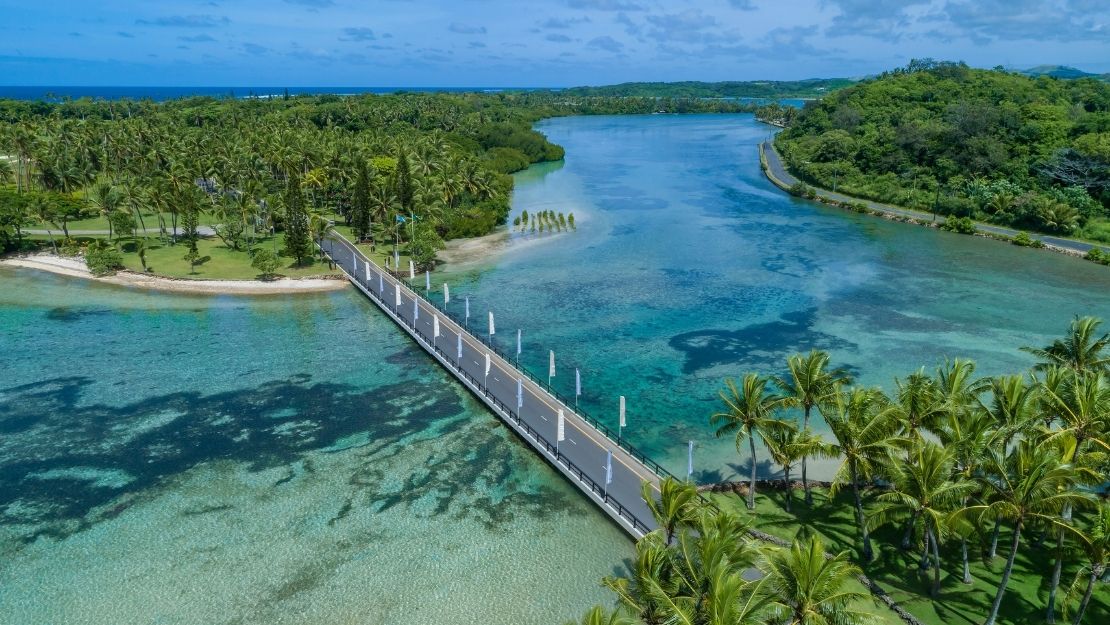 Aerial view of a modern bridge crossing a clear turquoise lagoon, surrounded by lush green palm trees and tropical foliage under a blue sky.