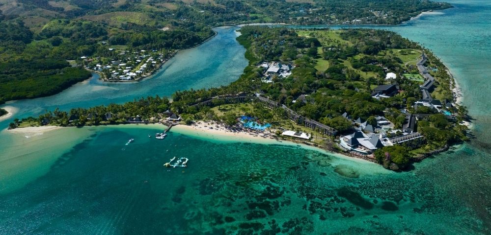 Aerial view of a tropical resort complex with buildings, pools, and a sandy beach along a turquoise bay.