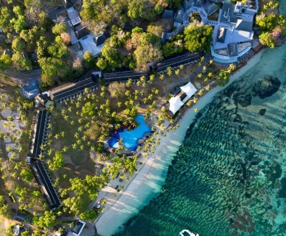 Aerial view of a tropical resort with a bright blue swimming pool, white sandy beach, lush green palm trees, and clear turquoise ocean water.