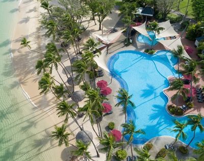 Aerial view of a tropical resort with a large, freeform swimming pool bordered by a sandy beach and lush palm trees.