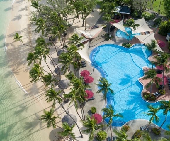 Aerial view of a tropical resort with a large, freeform swimming pool bordered by a sandy beach and lush palm trees.