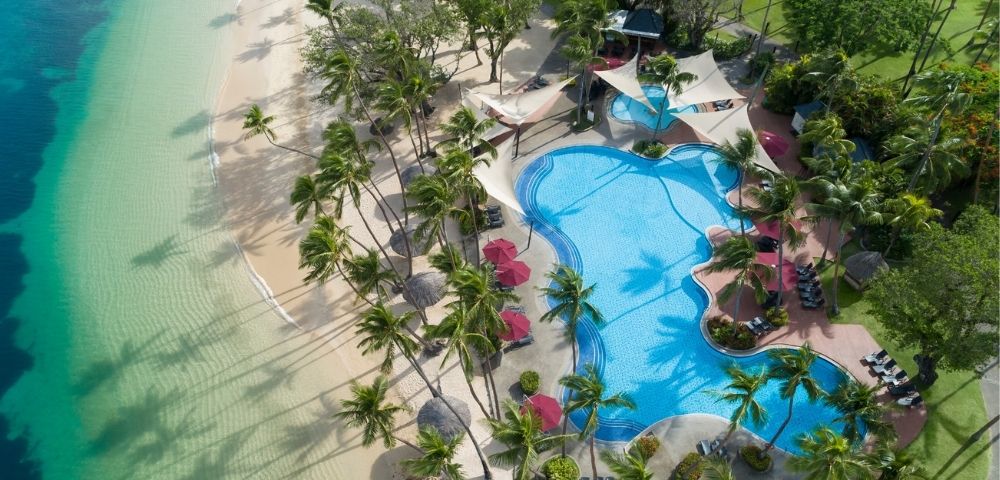 Aerial view of a tropical resort with a large, freeform swimming pool bordered by a sandy beach and lush palm trees.