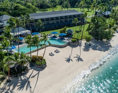 Aerial view of a tropical resort with a beachfront hotel, curved swimming pool, lounge chairs, palm trees, and a sandy beach meeting clear turquoise water.