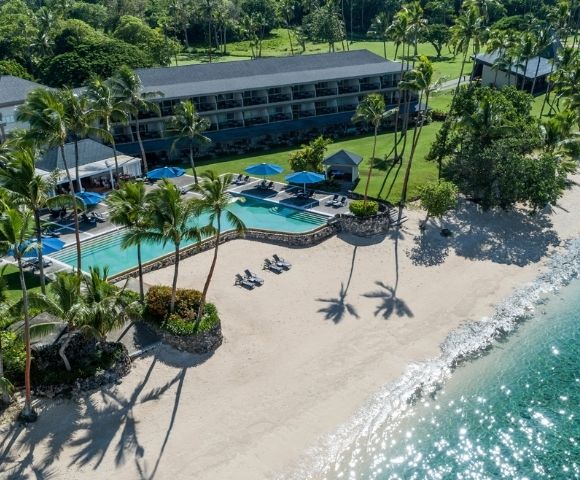 Aerial view of a tropical resort with a beachfront hotel, curved swimming pool, lounge chairs, palm trees, and a sandy beach meeting clear turquoise water.