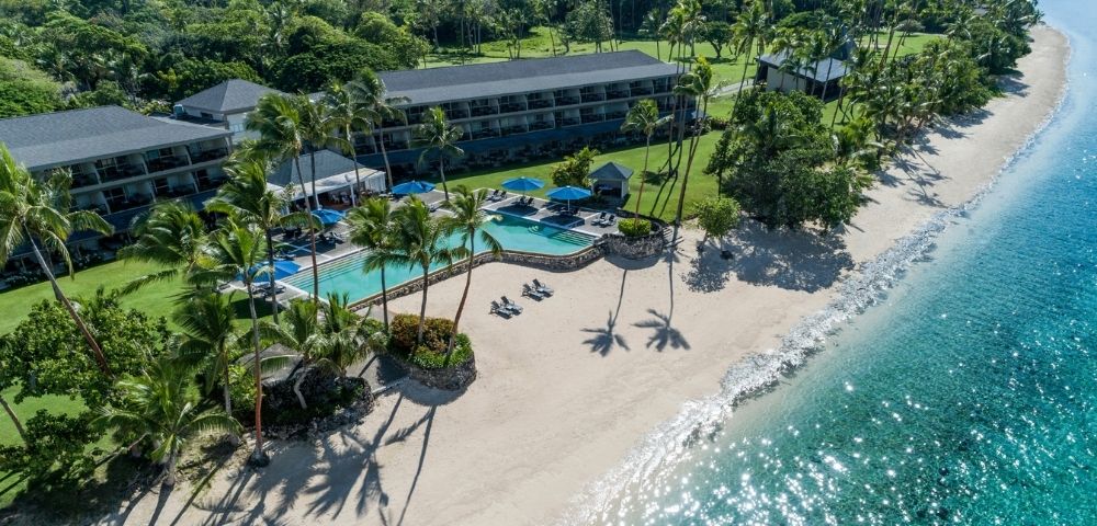 Aerial view of a tropical resort with a beachfront hotel, curved swimming pool, lounge chairs, palm trees, and a sandy beach meeting clear turquoise water.