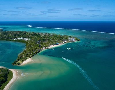 Aerial view of a lush, tropical island peninsula with white sand beaches, palm trees, and resort buildings, surrounded by clear turquoise and deep blue ocean waters. A boat leaves a white wake as it speeds across the water.