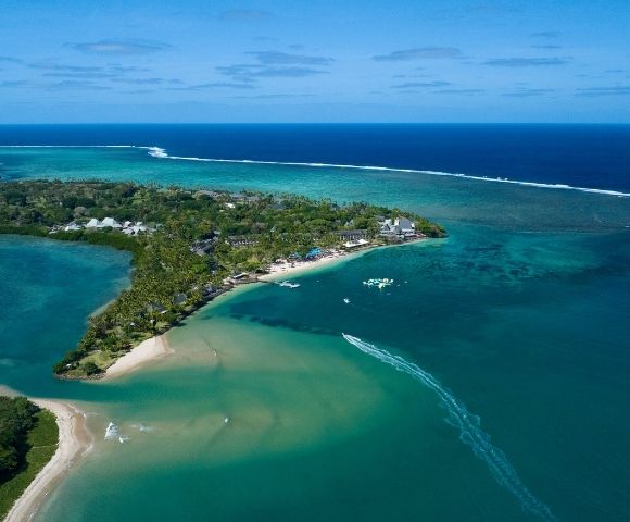 Aerial view of a lush, tropical island peninsula with white sand beaches, palm trees, and resort buildings, surrounded by clear turquoise and deep blue ocean waters. A boat leaves a white wake as it speeds across the water.