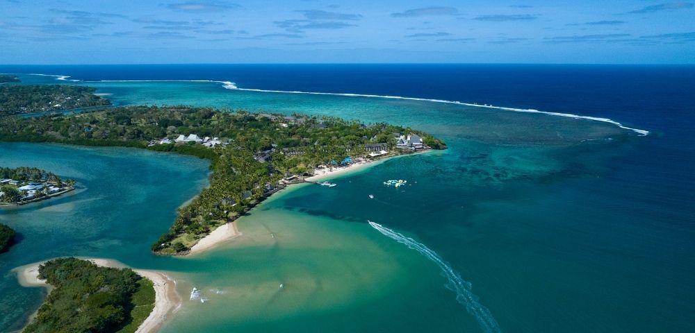 Aerial view of a lush, tropical island peninsula with white sand beaches, palm trees, and resort buildings, surrounded by clear turquoise and deep blue ocean waters. A boat leaves a white wake as it speeds across the water.