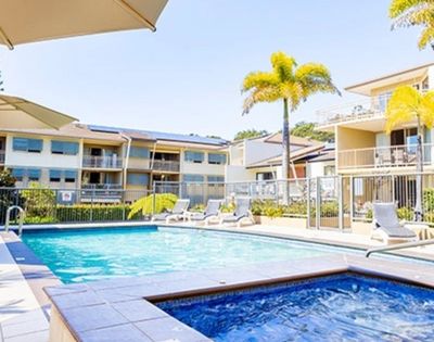 A resort-style swimming pool and hot tub are surrounded by lounge chairs and palm trees, with apartment buildings in the background under a clear blue sky.