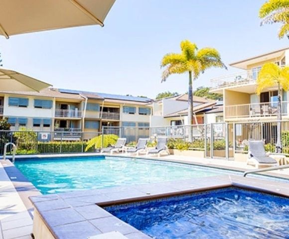 A resort-style swimming pool and hot tub are surrounded by lounge chairs and palm trees, with apartment buildings in the background under a clear blue sky.